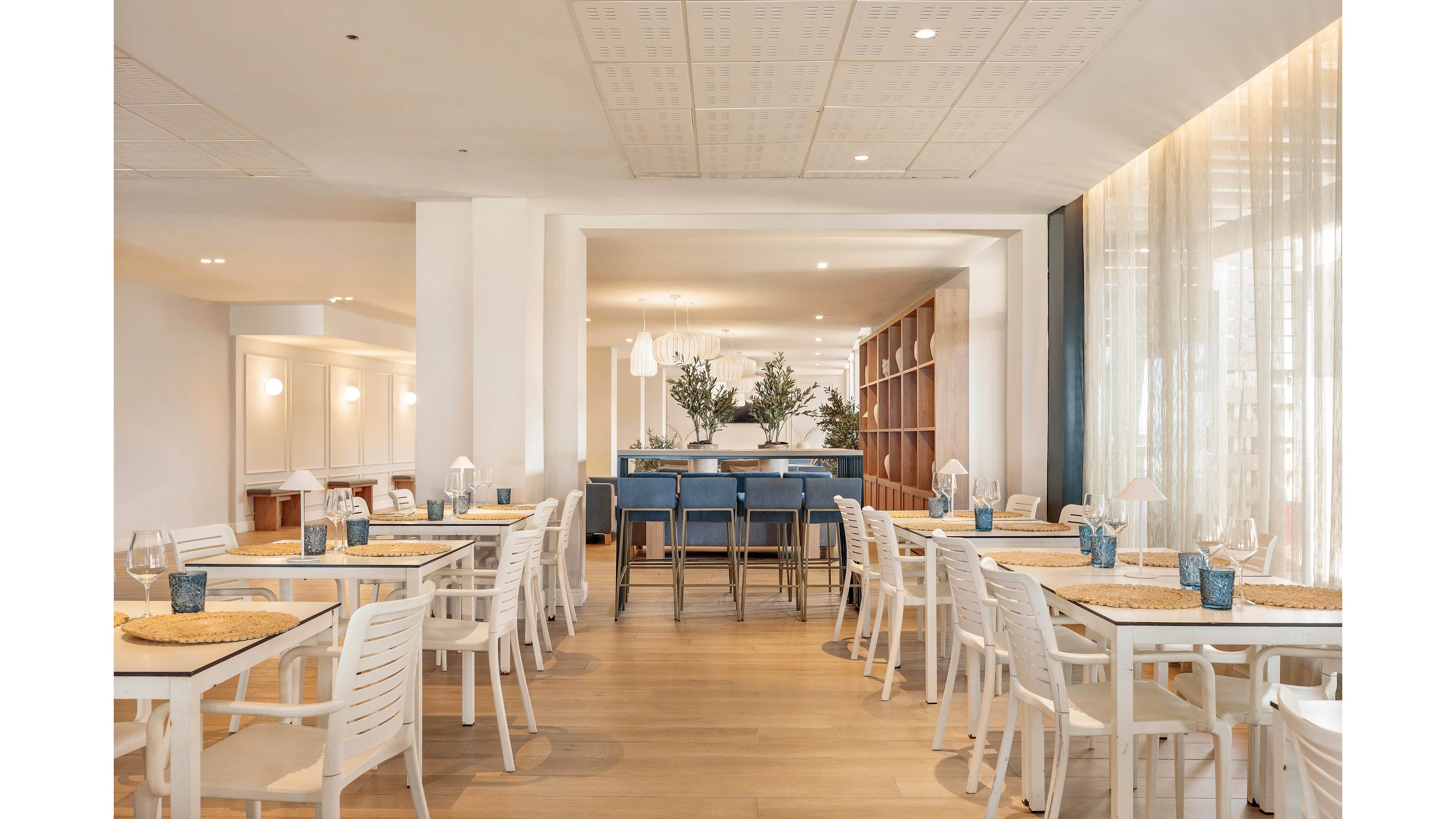 A bright modern restaurant dining room features neatly arranged white tables and chairs. Woven placemats and blue glass tumblers add subtle color accents to the neutral palette. In the background, a bar area with blue upholstered stools and decorative plants creates a focal point. Soft natural light filters through sheer curtains, giving the space an airy, inviting atmosphere.
