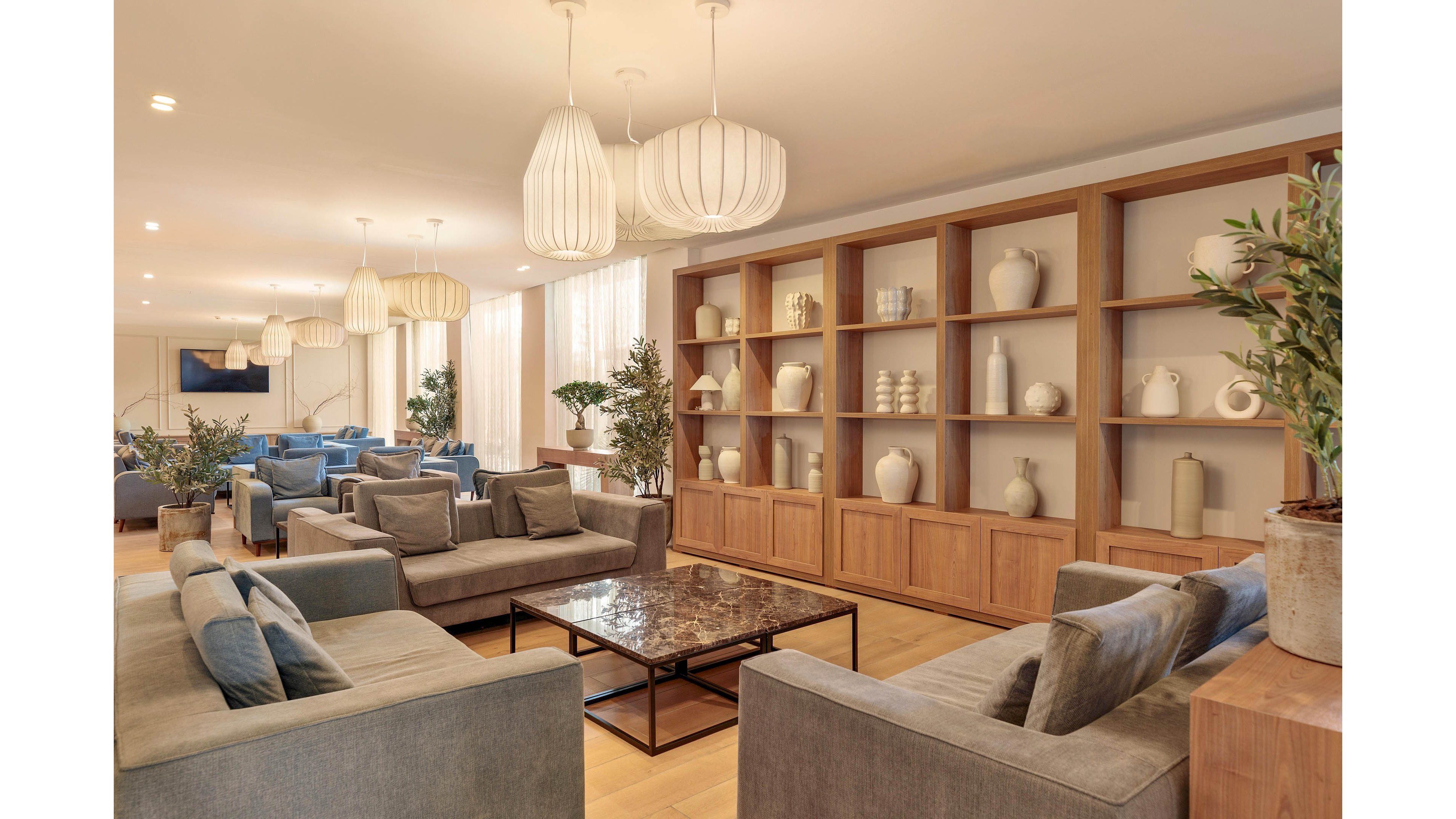 A spacious modern lounge features multiple grey sofas arranged around a central marble coffee table. Floor-to-ceiling wooden shelving along one wall displays decorative vases and ceramics. Large pendant lights hang from the ceiling, casting warm light over the room. Soft neutral tones, wooden flooring, and indoor plants create a calm, contemporary atmosphere.