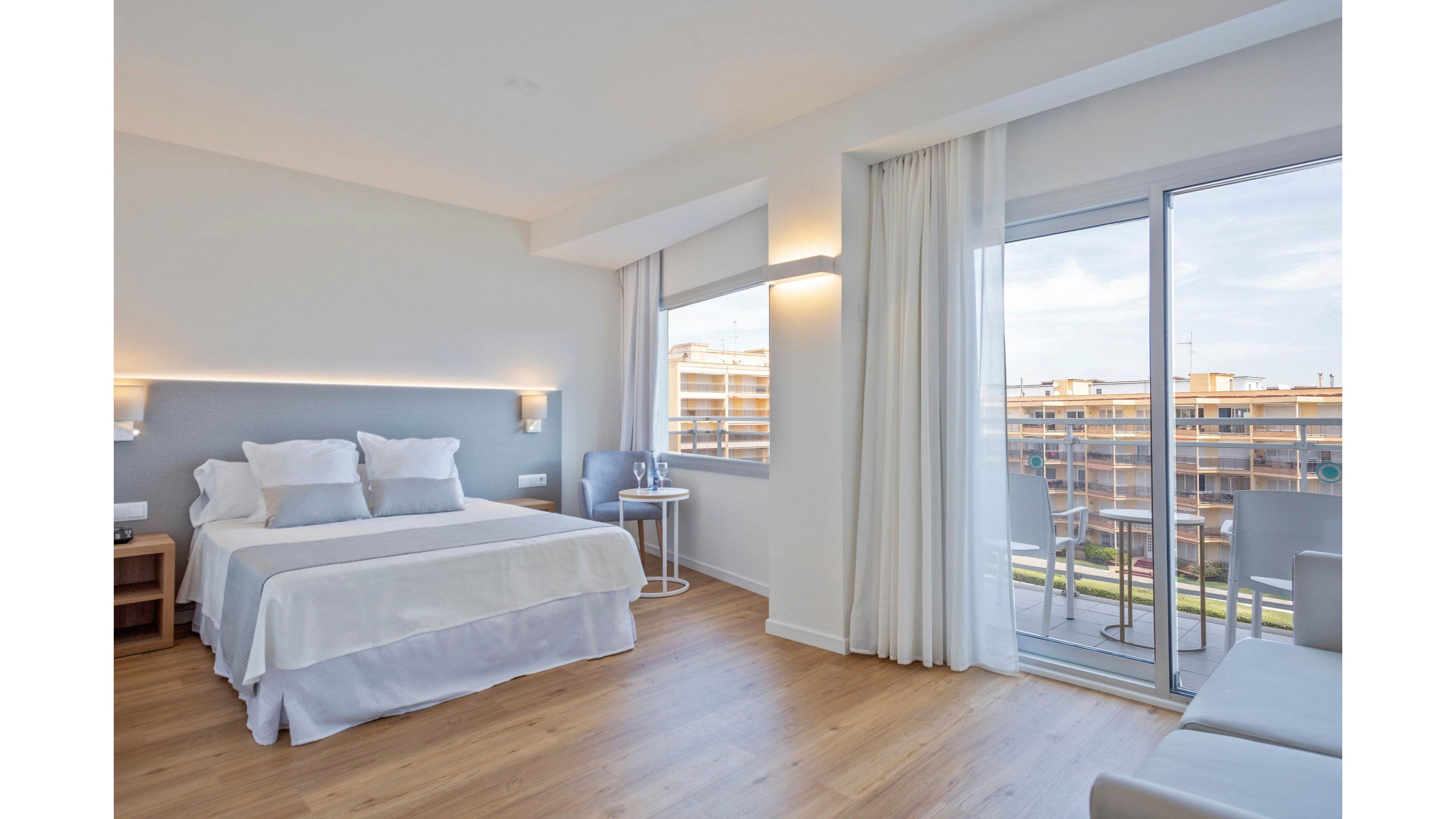 A contemporary hotel room features a neatly made double bed with white linens and multiple pillows against a light gray accent wall. The space includes wooden flooring, a small bedside table, and a round side table with two chairs near the window. Large floor-to-ceiling glass doors open to a balcony overlooking neighboring buildings under a bright sky. Soft white curtains and minimalist wall lighting create a clean, airy, and inviting atmosphere.