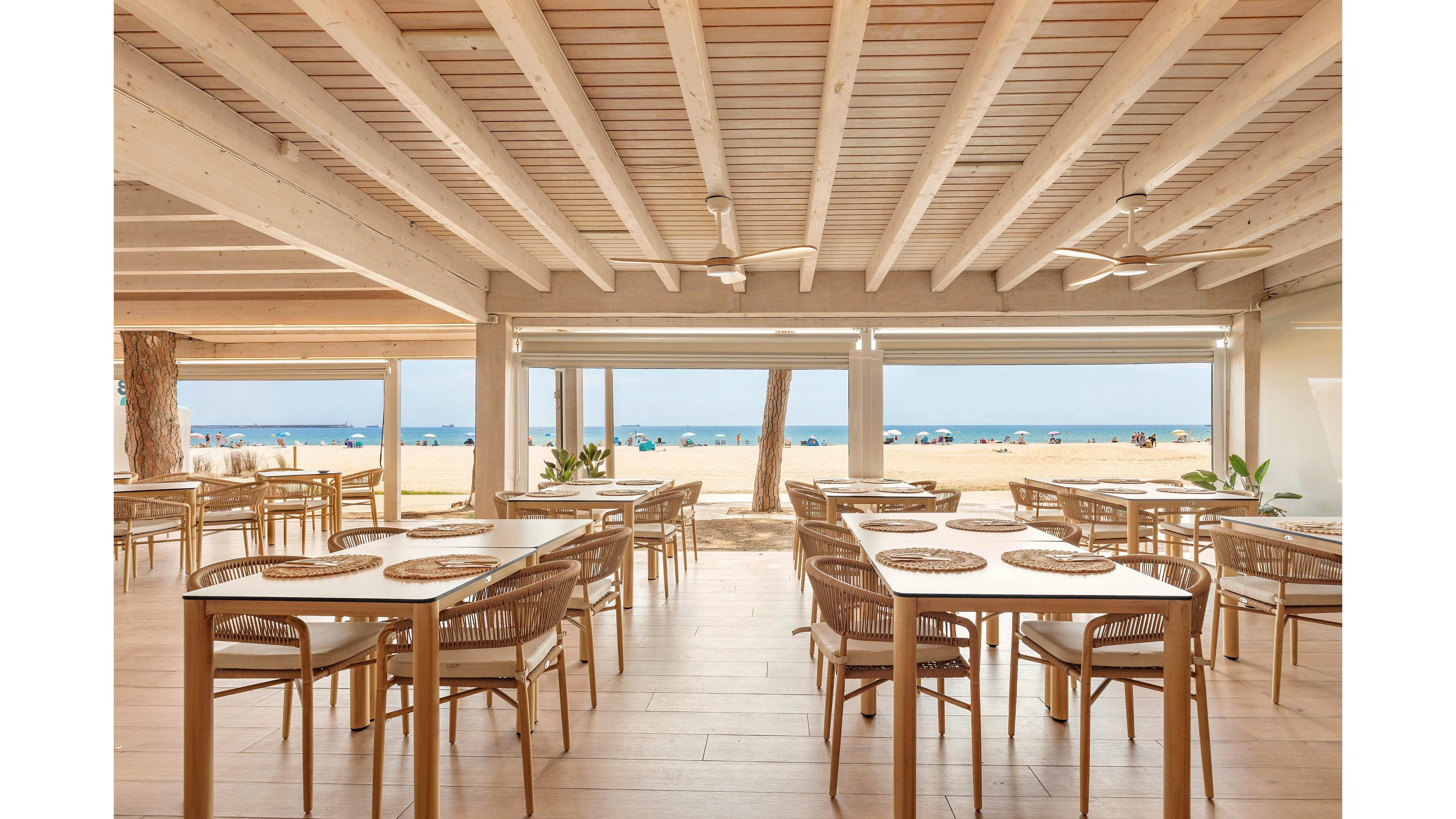 An airy beachfront restaurant features neatly arranged wooden tables and wicker chairs under a light wooden ceiling. Large open walls frame a clear view of the sandy beach and calm blue sea beyond. Ceiling fans and natural tones create a relaxed, coastal atmosphere. The space appears ready for guests, with placemats set on each table and bright daylight filling the interior.