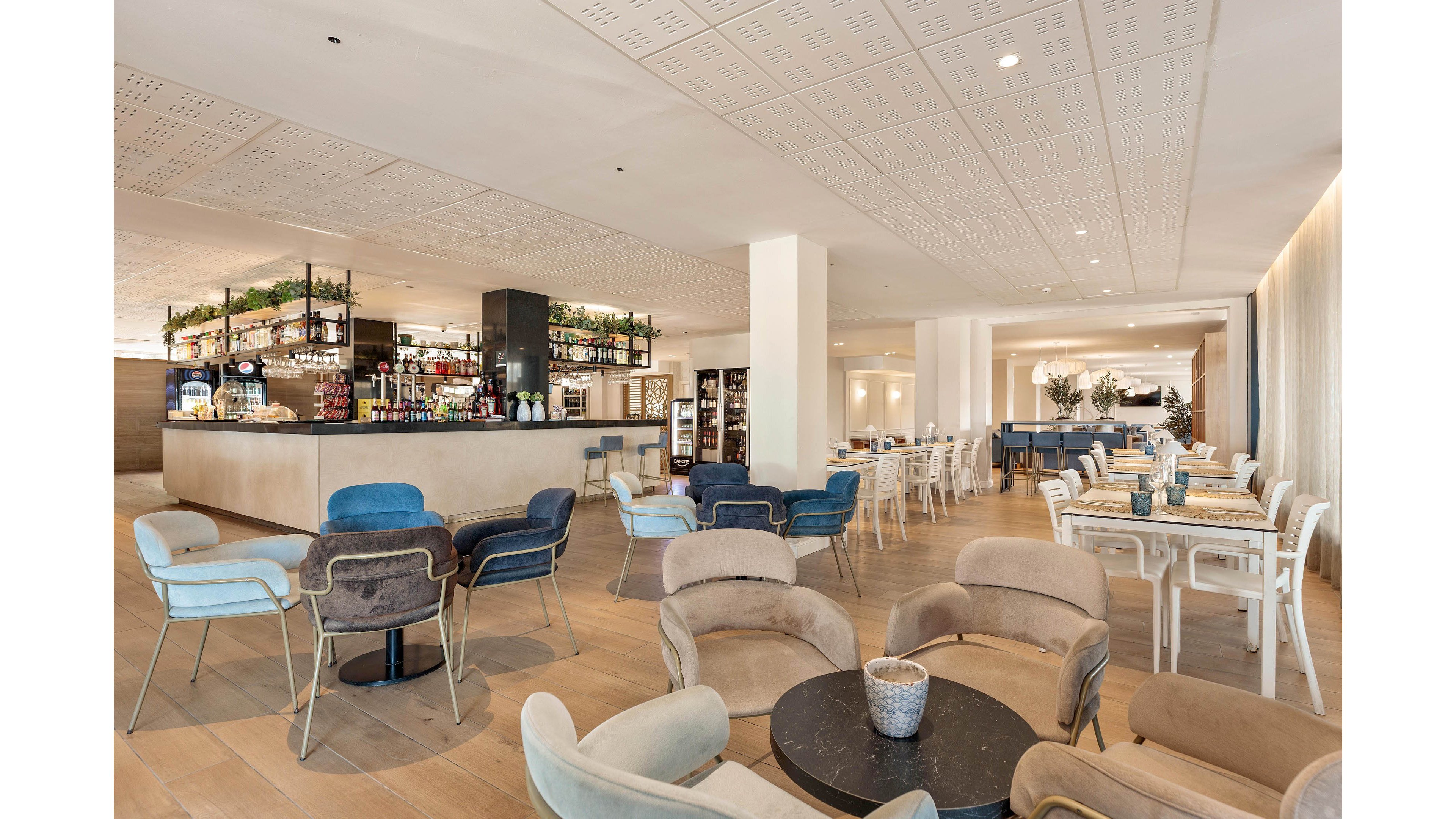 Spacious contemporary lounge and dining area featuring a central bar with bottles and glassware on display. Soft upholstered chairs and minimalist tables are arranged throughout the room, creating an inviting seating layout. Neutral tones with light wood flooring and white walls give the space an airy, modern feel. Large windows and subtle lighting enhance the relaxed, upscale atmosphere of the interior.