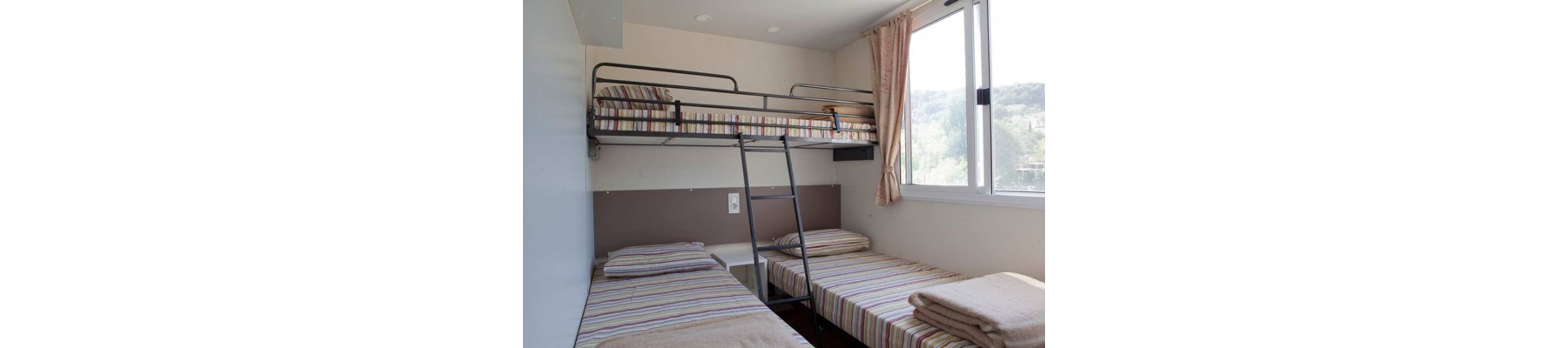 A compact bedroom features a metal bunk bed above two single beds arranged in an L-shape. All three beds have matching striped bedding and folded blankets at the foot. A ladder leads to the upper bunk, and a large window with curtains lets in natural light and a view of greenery outside. The room has a simple, functional design suitable for shared accommodation or a hostel.