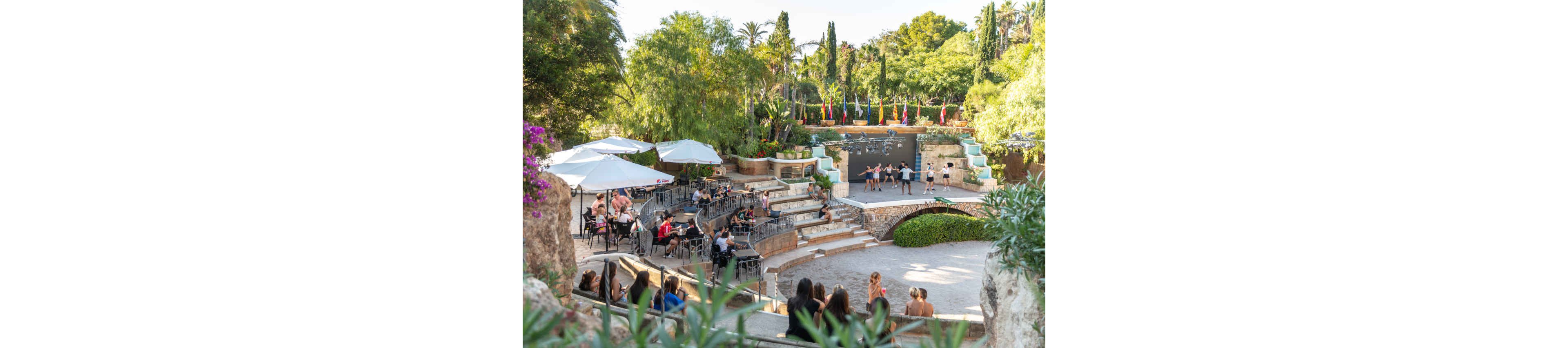 An outdoor amphitheater hosts a live performance on a small stage surrounded by greenery. Audience members sit at café-style tables under white umbrellas and on stone steps, watching dancers or entertainers. Colorful flags line the back of the stage, adding a festive touch to the scene. Tall trees, flowering plants, and bright daylight create a relaxed, vacation-like atmosphere.