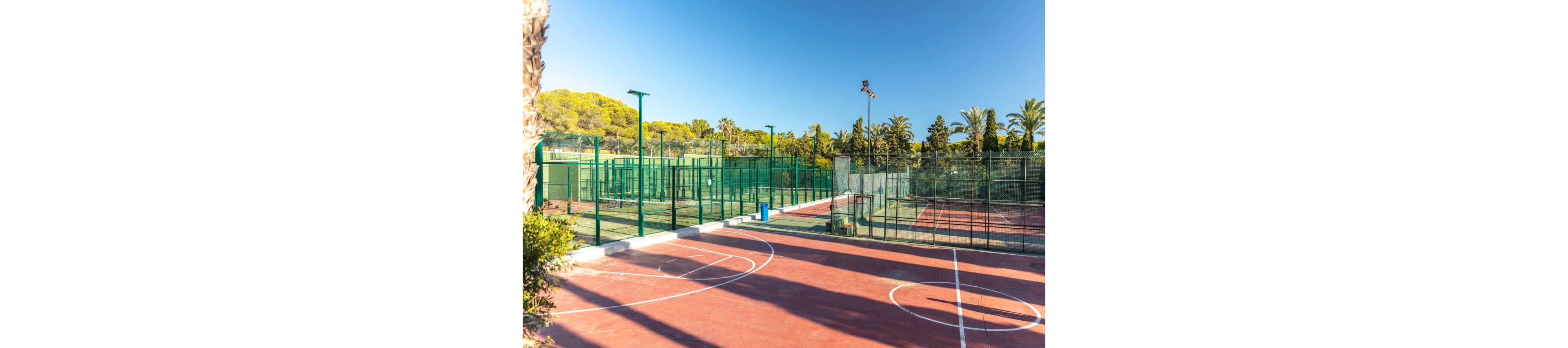 An empty outdoor sports complex features multiple fenced courts under a clear blue sky. A red basketball or multi-sport court with white lines is visible in the foreground. Behind it, several green tennis or paddle courts are enclosed by tall metal fences. Palm trees and lush greenery surround the area, creating a bright and relaxed atmosphere.