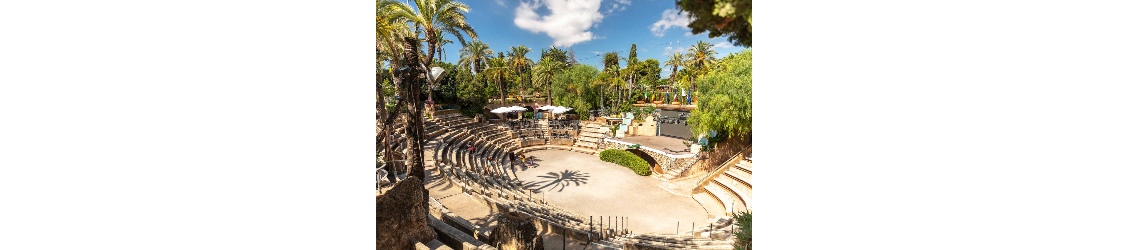 An open-air amphitheater with curved stone seating is nestled among tall palm trees and dense greenery. A few visitors are scattered across the terraces, emphasizing the scale of the venue. Sunlight casts strong shadows onto the central performance area, highlighting the circular design. In the background, colorful structures and umbrellas suggest a nearby park or leisure attraction.