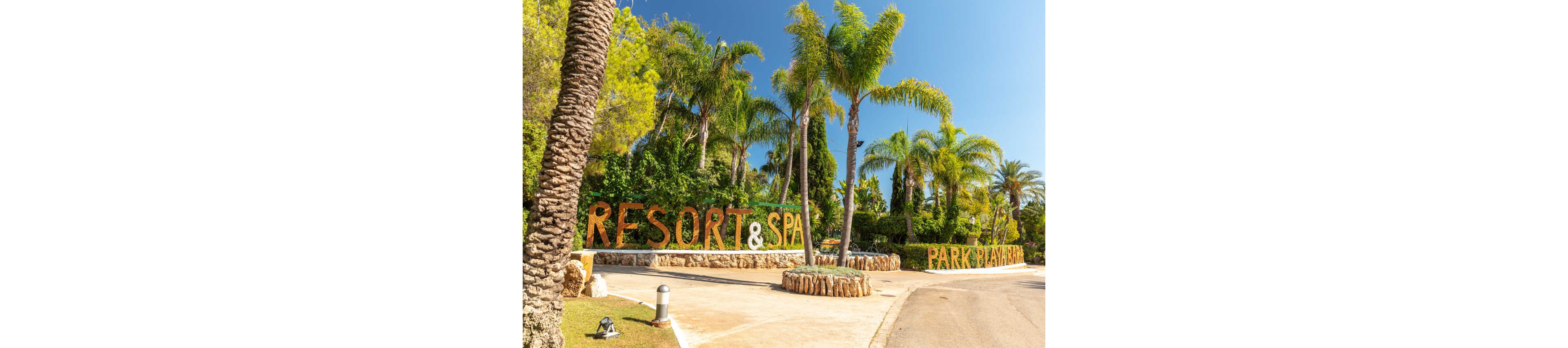 A sunny entrance to a resort and spa is framed by tall palm trees and lush greenery. Large letters spelling out “RESORT & SPA” and “PARK PLAYA” are integrated into the landscaping along a curved driveway. The scene suggests a warm, tropical climate with clear blue skies and bright daylight. Stone walls, decorative rocks, and pathway lighting enhance the welcoming atmosphere.