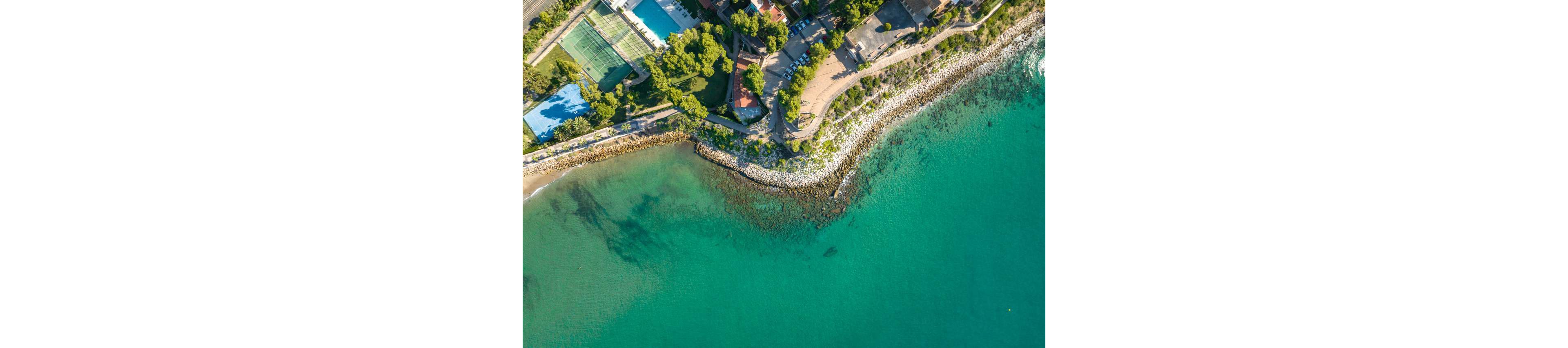 A drone captures an aerial view of a rocky shoreline meeting clear turquoise water. A curved coastal road and walking path follow the edge of the land, bordered by trees and greenery. Sports courts and buildings are visible among the trees near the top of the image. Gentle waves wash against the rocks, creating a tranquil seaside atmosphere.
