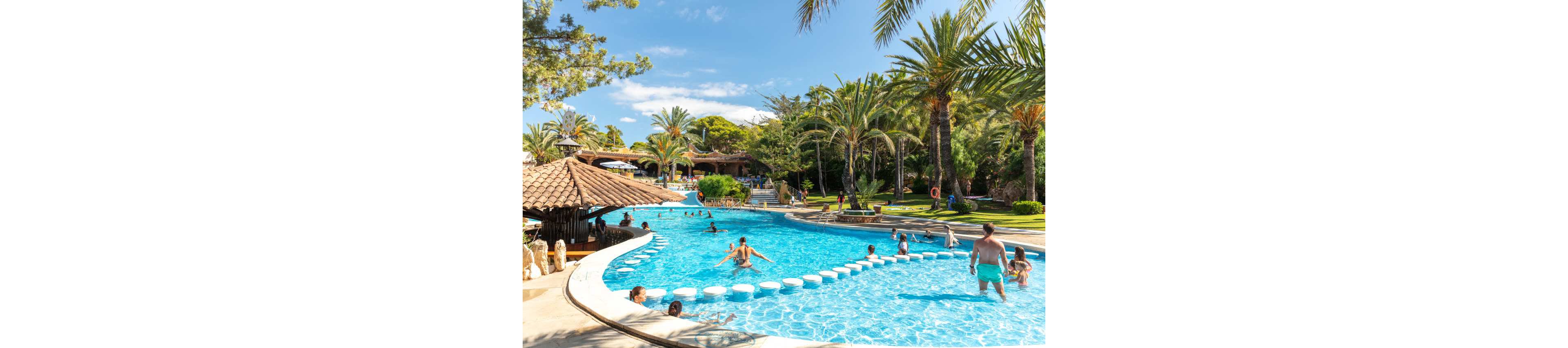 A large outdoor swimming pool curves through a lush resort surrounded by palm trees and green lawns. Families and children play in the clear blue water, some walking across circular stepping stones. Sunbathers relax on loungers under umbrellas while others wade near the pool edge. The bright daylight and tropical landscaping create a cheerful, vacation atmosphere.