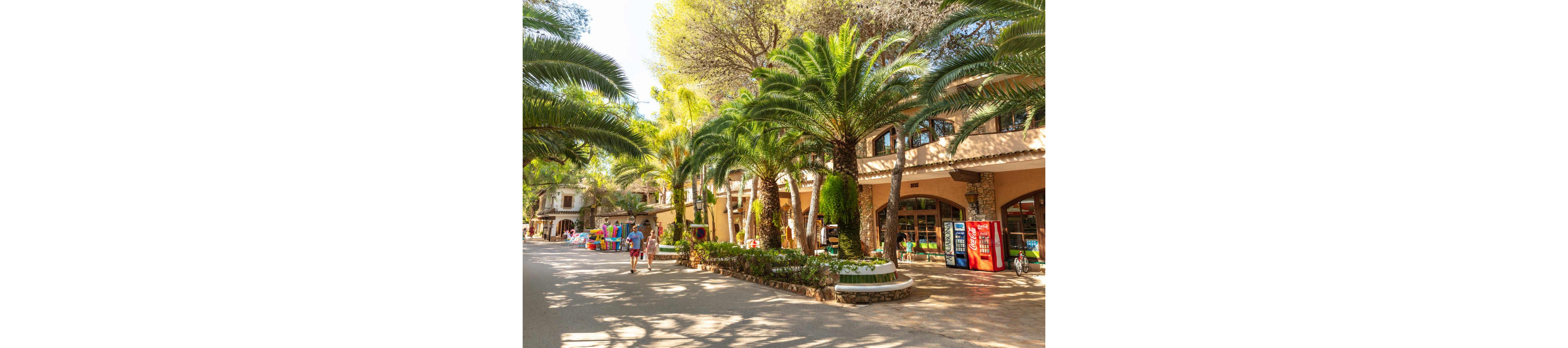 A sunlit pedestrian street is lined with tall palm trees and Mediterranean-style buildings. People stroll casually along the wide pathway, suggesting a relaxed vacation atmosphere. Colorful vending machines and shopfronts appear on the right side, while children’s rides or toys are visible in the distance. Warm light and lush greenery create a bright, inviting holiday scene.
