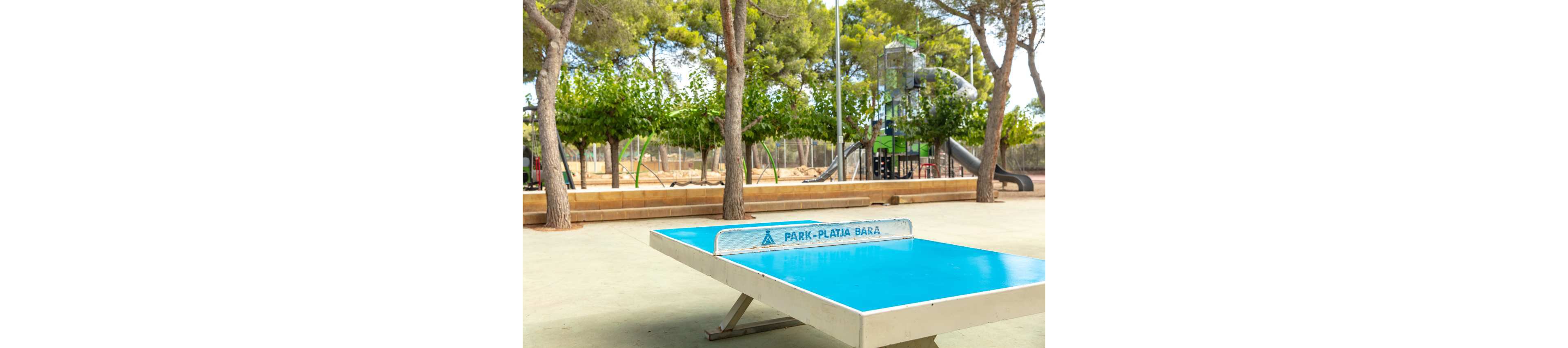 A bright blue outdoor ping pong table stands in the foreground of a park. Behind it, a children’s playground with slides and climbing structures is surrounded by trees. The setting appears calm and sunny, with green foliage providing shade. The table net shows the text "PARK-PLATJA BARA" clearly printed on it.