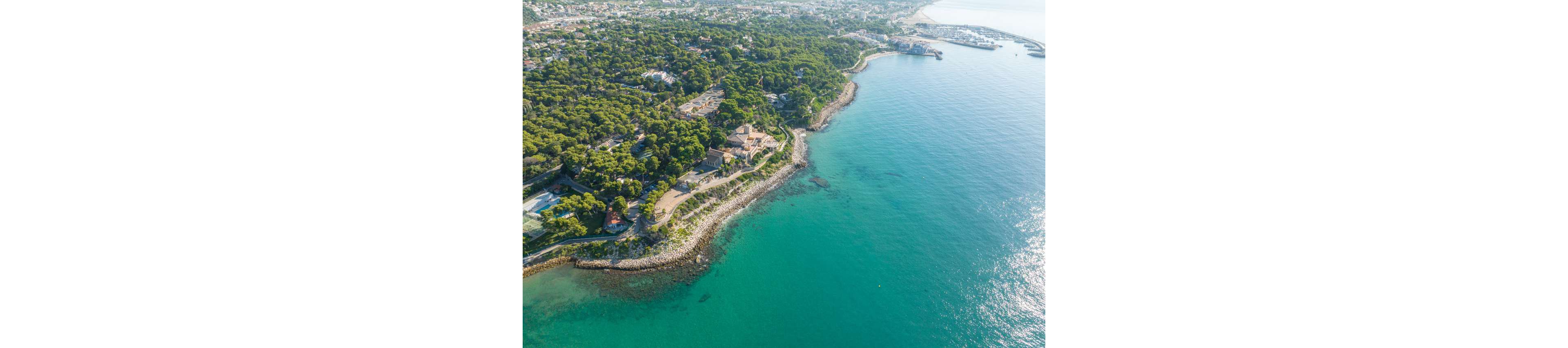 High-angle aerial photography captures a lush green coastline meeting clear turquoise sea. A rocky shoreline curves gently along the water, with trees and vegetation covering most of the land. Buildings and villas are nestled among the forested area, and a distant town stretches further inland. The calm water and bright daylight create a serene, summery atmosphere.
