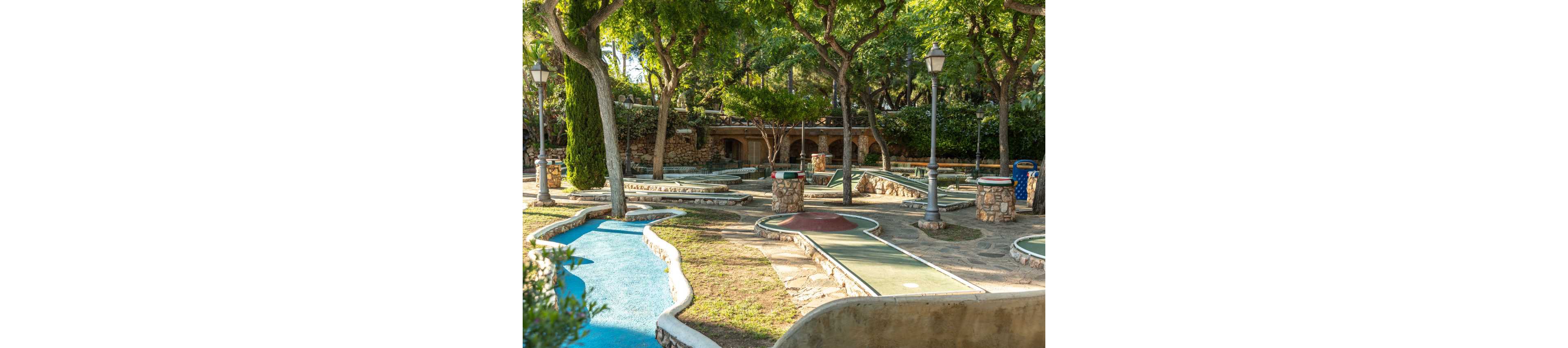 An empty mini golf course winds through a shaded outdoor park. Curved putting lanes with green and red surfaces are bordered by low stone walls and concrete edges. Tall trees, lampposts, and rustic stone structures create a relaxed, natural setting. Warm sunlight and long shadows suggest a peaceful afternoon atmosphere.