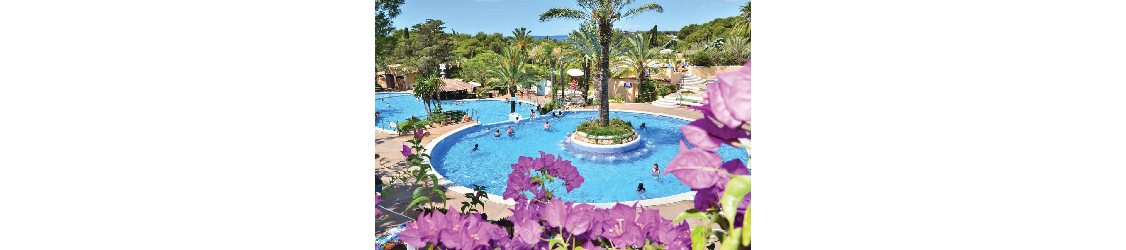 A large curving swimming pool is surrounded by lush palm trees and flowering plants at a sunny resort. People of various ages relax and play in the clear blue water and on the poolside deck. Additional pools and waterslides are visible in the background, creating a family-friendly water park atmosphere. Bright purple flowers frame the foreground, adding vibrant color to the tropical holiday scene.