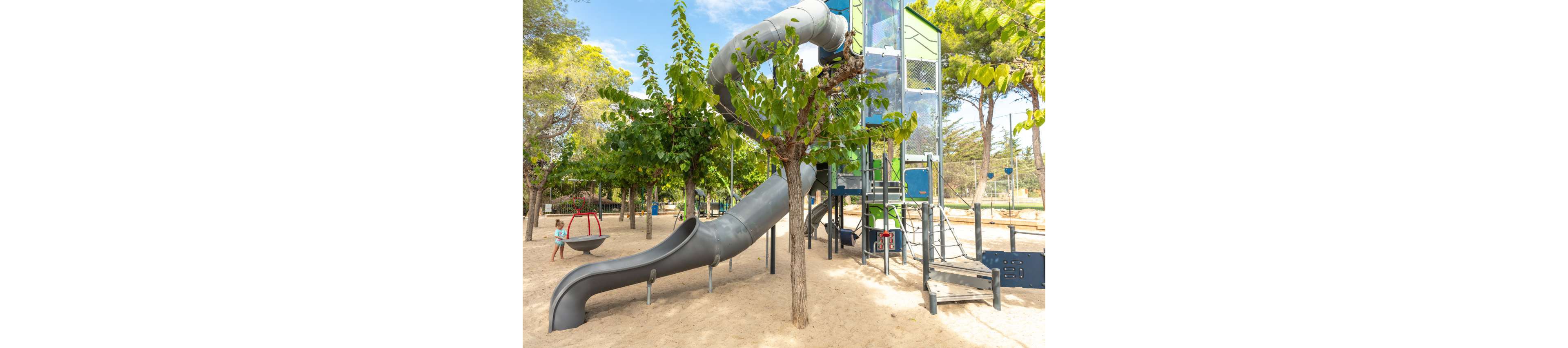 A large outdoor playground features a tall climbing tower connected to a winding enclosed tube slide and an open slide. The structure stands on a sandy surface surrounded by several young trees providing partial shade. In the background, a child plays near a round swing while other play equipment and fencing are visible. The scene is bright and sunny, suggesting a warm day in a family-friendly park.