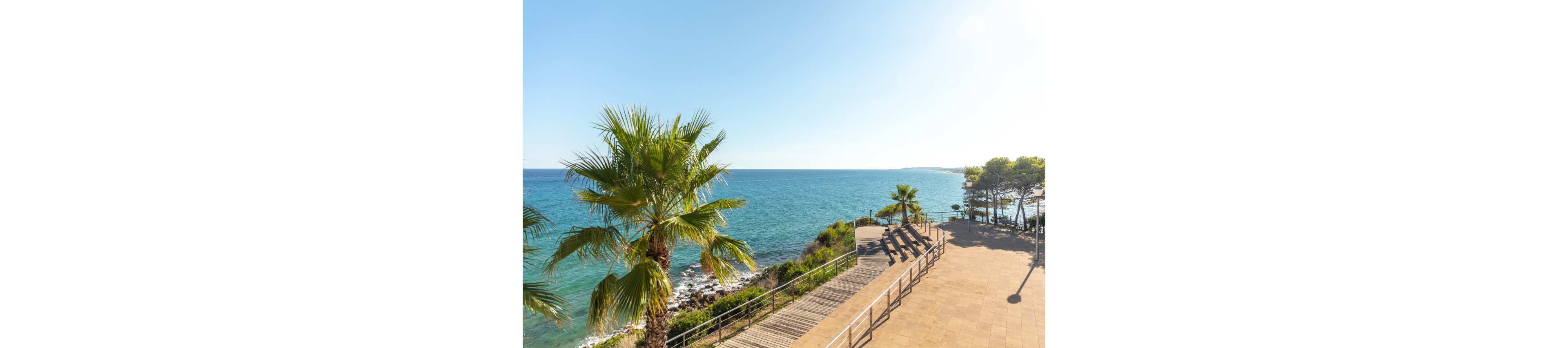 A bright coastal scene shows a sunlit promenade overlooking a calm blue sea. Palm trees line the wooden boardwalk that runs along the rocky shoreline. Several empty lounge chairs face the water, suggesting a relaxing seaside retreat. The clear sky and strong sunlight create a warm, inviting holiday atmosphere.
