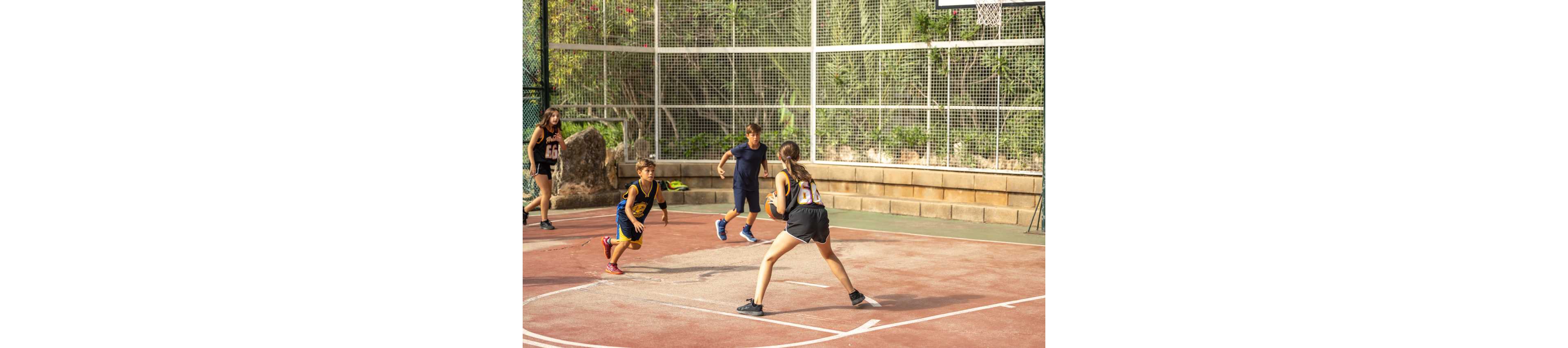 A group of children play a casual basketball game on an outdoor court. One girl in a numbered jersey dribbles the ball while other kids move to defend and support. The court is surrounded by a tall wire fence and greenery in the background. Warm sunlight creates long shadows on the red and white court surface.