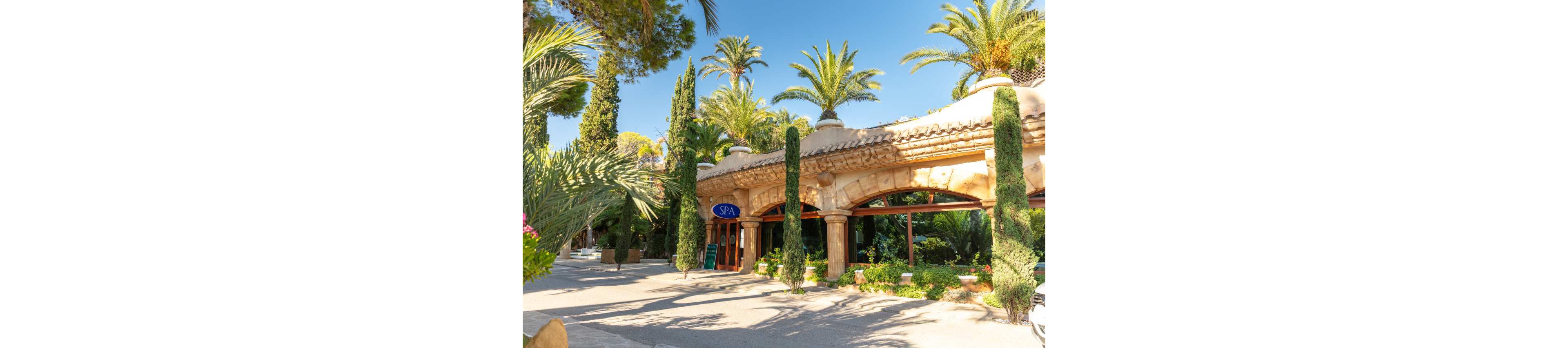 A Mediterranean-style spa building is surrounded by tall palm trees and lush greenery. The structure features stone arches, terracotta roof tiles, and decorative columns along the facade. Bright sunlight casts warm shadows across the entrance area and pathway. A blue sign reading "SPA" is clearly visible above the doorway, indicating the wellness facility.