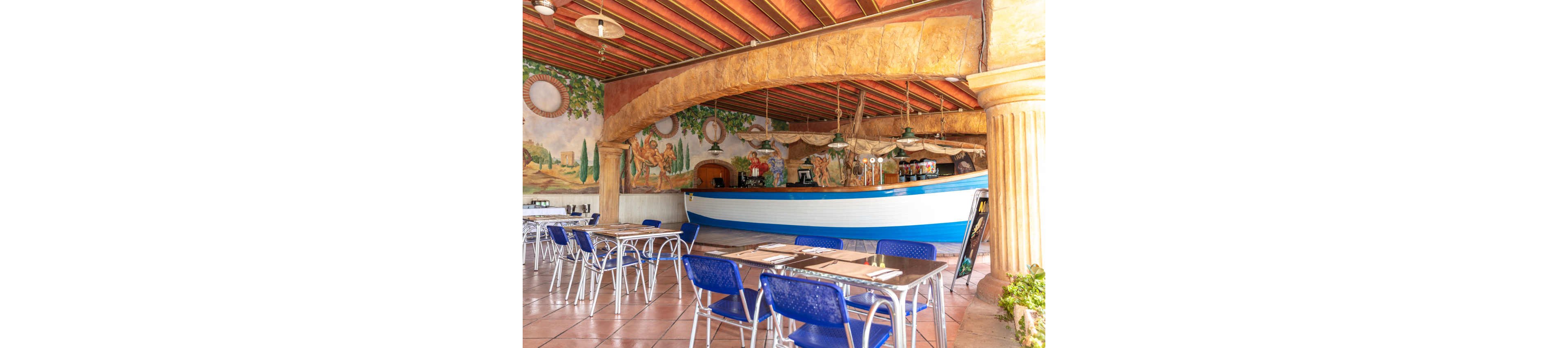 A bright restaurant interior features a large bar counter shaped like a white and blue boat. Blue metal chairs and simple tables with placemats are arranged across the tiled floor. The walls are decorated with painted murals of classical scenes and greenery, while rustic columns and a wooden ceiling enhance the Mediterranean feel. The space appears empty of people, emphasizing the decor and seating layout.