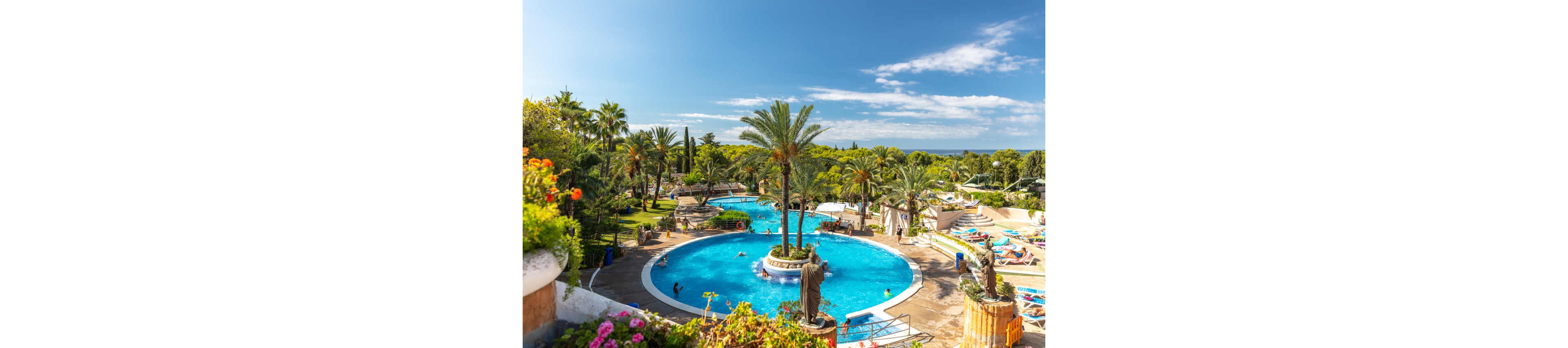 A large circular swimming pool is surrounded by palm trees and sun loungers at a tropical resort. Guests relax on deck chairs and walk along the poolside under bright sunshine. Lush green gardens and landscaped paths frame the water, creating a vacation atmosphere. In the distance, more pools and greenery extend toward the horizon under a clear blue sky.