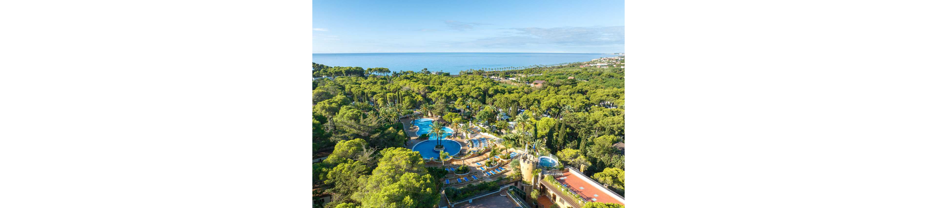 A lush coastal resort is surrounded by dense green trees and gardens. Multiple outdoor swimming pools with sun loungers and palm trees form the center of the complex. A tower-like building and terracotta rooftops add architectural interest in the foreground. In the distance, the calm blue sea stretches to the horizon under a clear sky.