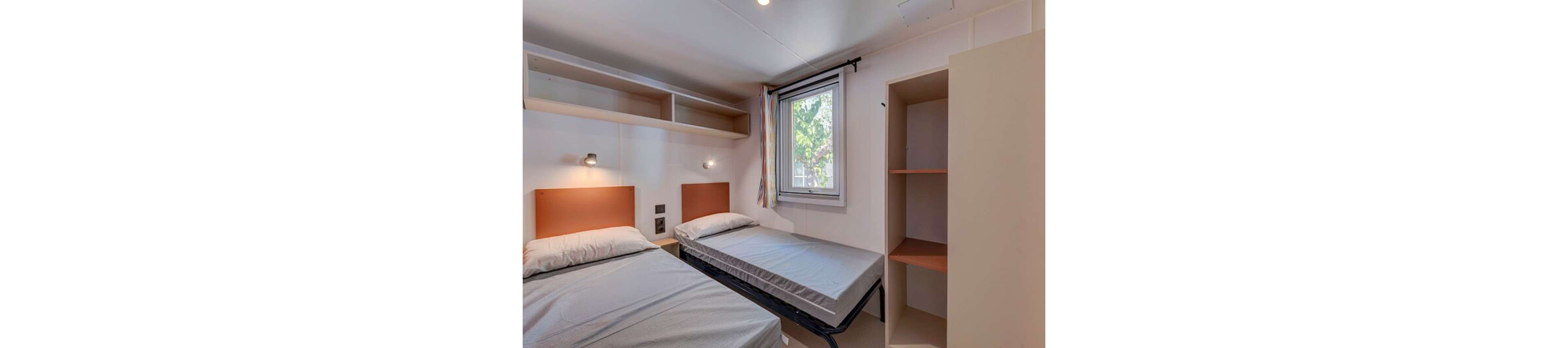 Interior view of a small bedroom with two single beds, one slightly pulled out. The room features a window with a view of greenery outside, allowing natural light to brighten the space. Open shelving above the beds and an empty open wardrobe provide storage options. The decor is simple and functional, with neutral walls and bedding creating a clean, minimalist look.