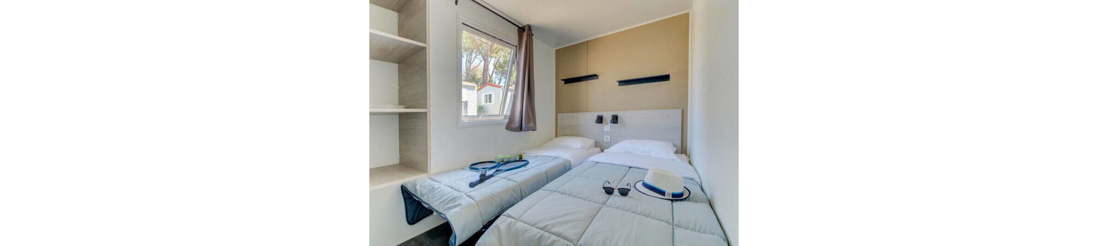 Compact modern bedroom with two single beds neatly made in light gray bedding. A window with a brown curtain lets in daylight and shows neighboring mobile homes outside. Tennis rackets and balls rest on one bed, while a hat and sunglasses are placed on the other, suggesting a vacation or resort stay. Minimalist shelves, wall-mounted reading lights, and neutral walls create a clean, relaxed atmosphere.