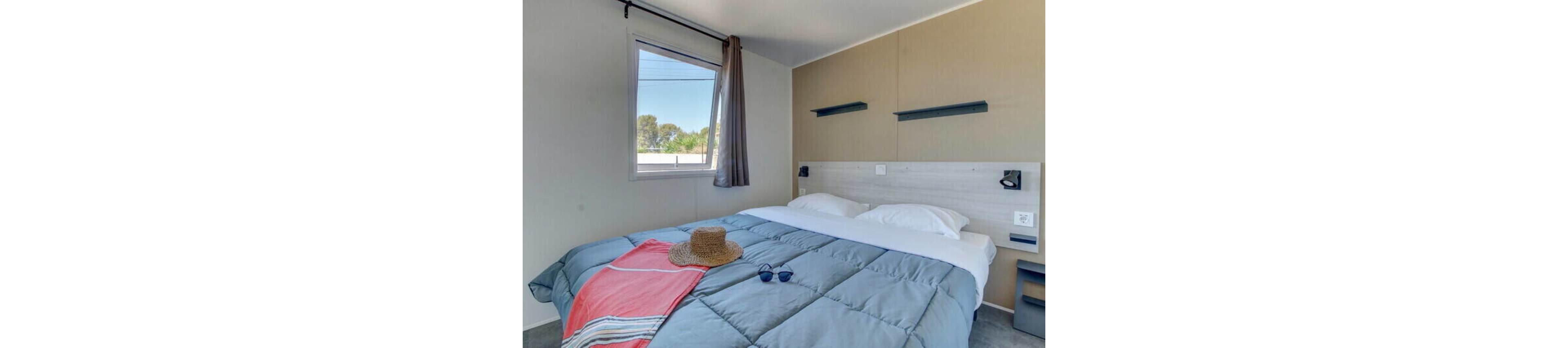 A neatly made double bed with a blue comforter and white pillows is the focal point of this modern bedroom. A straw hat and sunglasses rest on a coral and gray striped towel at the foot of the bed, suggesting a relaxed vacation setting. Natural light enters through a window with sheer and dark curtains, revealing trees and sky outside. Minimalist wall shelves and bedside fixtures contribute to the clean, contemporary style of the room.