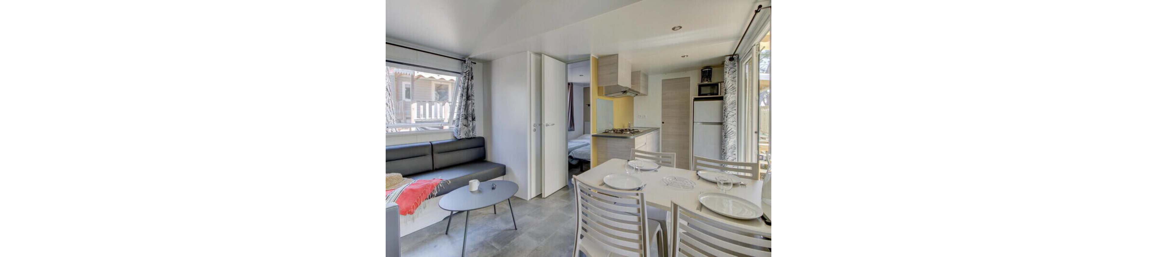 Bright, compact apartment interior featuring an open-plan living and dining area. A small kitchen with stove and fridge stands beside a set table with white plates and glasses. A gray sofa with a red throw and coffee table sits near large windows with patterned curtains. A partially open door reveals a cozy bedroom, suggesting a vacation rental or mobile home setting.
