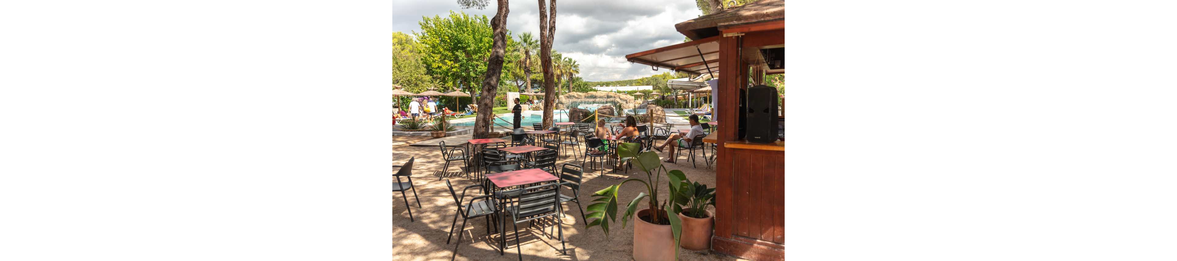 An outdoor café area with metal tables and chairs overlooks a swimming pool. A few people in swimwear relax near the water under umbrellas and trees. The scene is set on a sunny day with scattered clouds and lush greenery in the background. A wooden snack bar and potted plants frame the foreground, creating a casual holiday atmosphere.