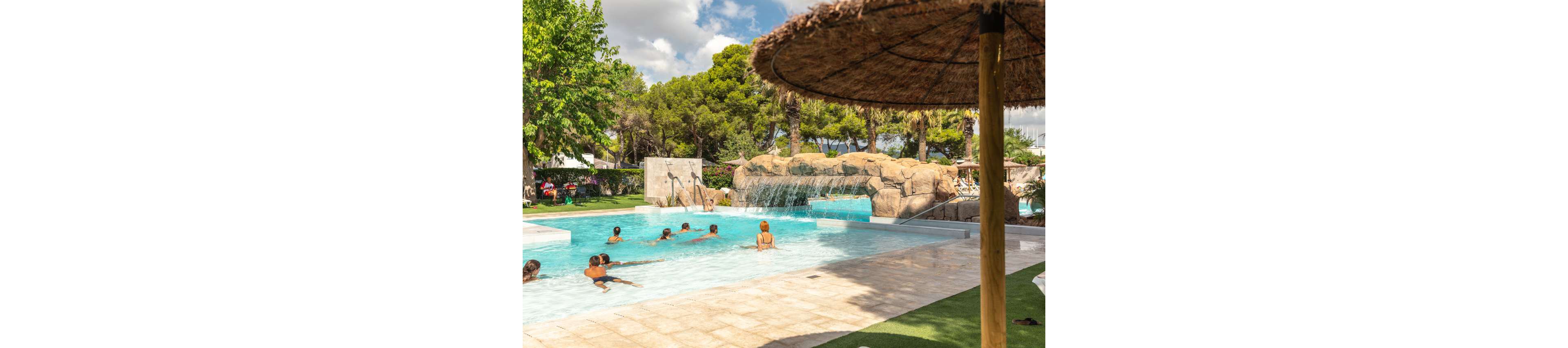 Several people enjoy a large outdoor swimming pool with a rock waterfall feature. The scene is set at a sunny resort surrounded by lush green trees and palm trees. Adults and children wade and swim in the clear blue water near the shallow edge. A thatched umbrella and paved pool deck create a relaxed vacation atmosphere under a partly cloudy sky.