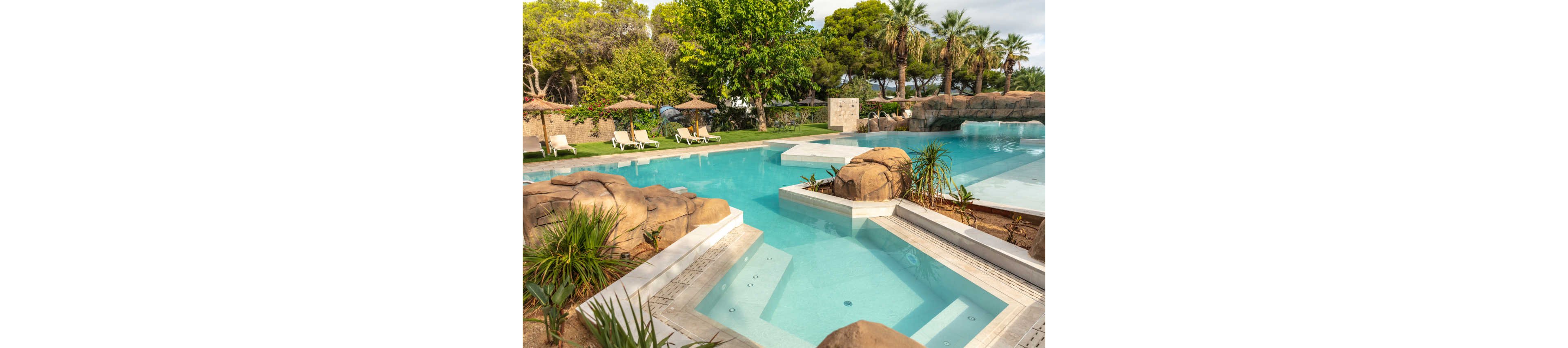 A large resort-style swimming pool features clear turquoise water and integrated rock formations. In the foreground, a shallow spa or wading area steps down into the main pool. Sun loungers with umbrellas are arranged on a grassy lawn beside the water. Lush trees and palm trees surround the pool, creating a tropical, relaxing atmosphere.