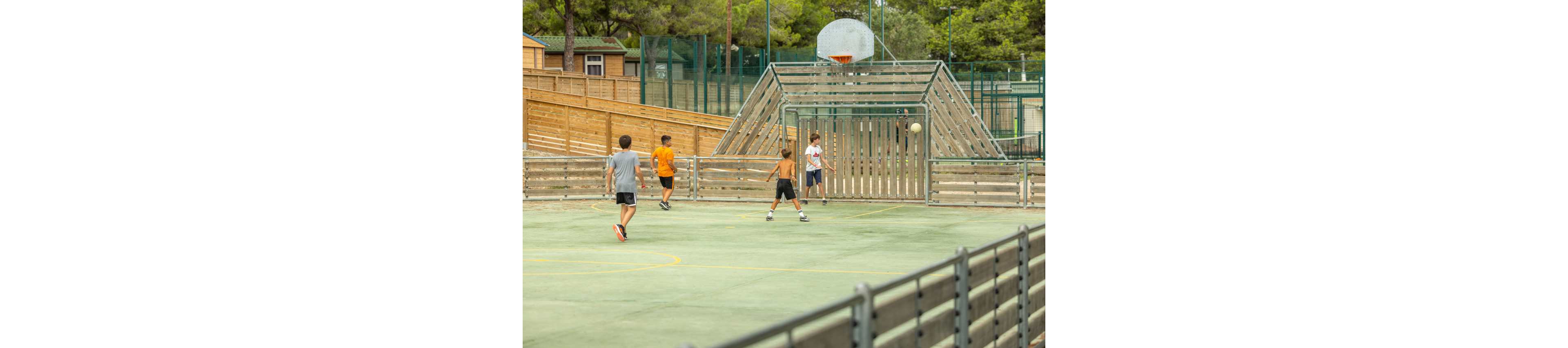 A group of teenage boys play soccer on an outdoor sports court. One player in an orange shirt runs toward the goal while another prepares to shoot the ball. The fenced court is surrounded by wooden barriers, metal railings, and tall trees in the background. The scene feels casual and energetic, suggesting a friendly game in a community or school setting.