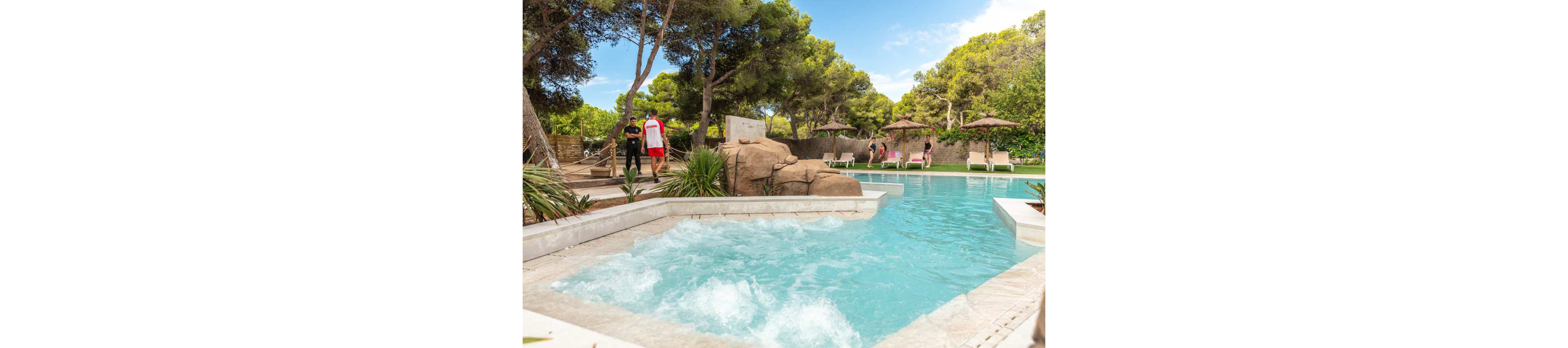 An outdoor resort-style pool features a bubbling jacuzzi section in the foreground. Guests relax on sun loungers and walk along the poolside deck surrounded by lush green trees. Natural rock formations and tropical plants enhance the tranquil atmosphere. The bright blue water and clear sky create a summery, vacation mood.