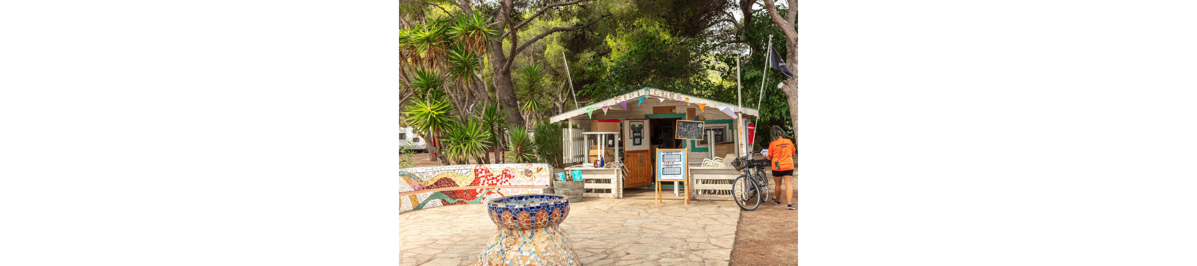 A small wooden café kiosk stands among tall trees in an outdoor campsite setting. Colorful bunting decorates the roofline, and chalkboard menus are displayed at the entrance. In the foreground, a mosaic-tiled fountain and low wall add vibrant artistic detail. A person in an orange shirt stands beside a bicycle near the kiosk, enhancing the relaxed holiday atmosphere.