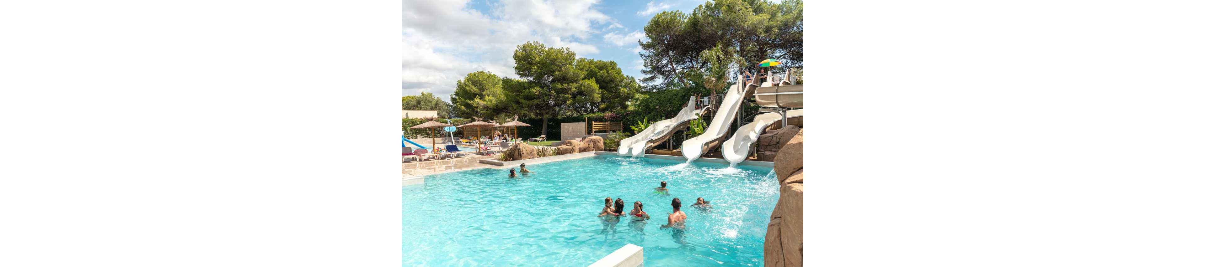 An outdoor swimming pool is filled with people enjoying a sunny day. Several children and adults play in the clear blue water while others relax on loungers under umbrellas. Large white water slides curve down into the pool from a rocky platform, adding a sense of fun and excitement. Lush green trees and landscaped rocks surround the pool area, creating a relaxed vacation atmosphere.