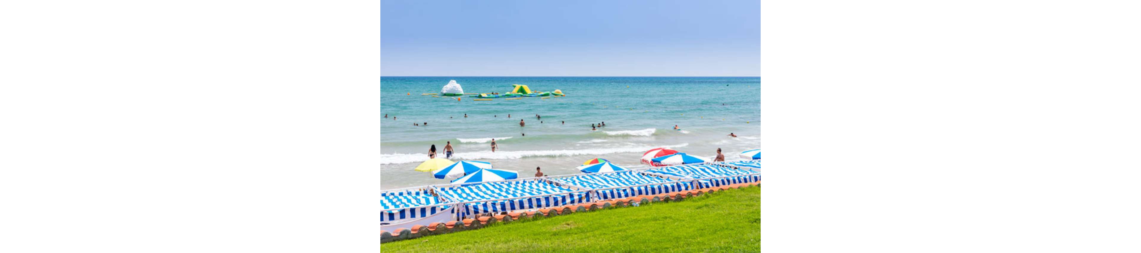 A sunny seaside beach is lined with rows of blue and white striped umbrellas and lounge chairs. People are swimming and playing in the shallow turquoise water near the shore. Farther out, large inflatable structures float on the waves, adding a playful element to the scene. A strip of bright green grass in the foreground contrasts with the sandy beach and calm blue sky.