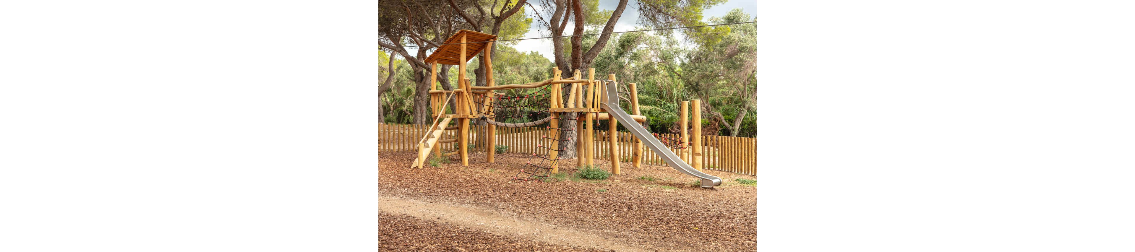 A large wooden playground structure stands in an outdoor park surrounded by trees. The play set features a metal slide, rope climbing nets, ladders, and elevated platforms. The ground is covered with wood chips, and a wooden fence encloses the area. Tall trees and green foliage create a natural, shaded backdrop for the play area.