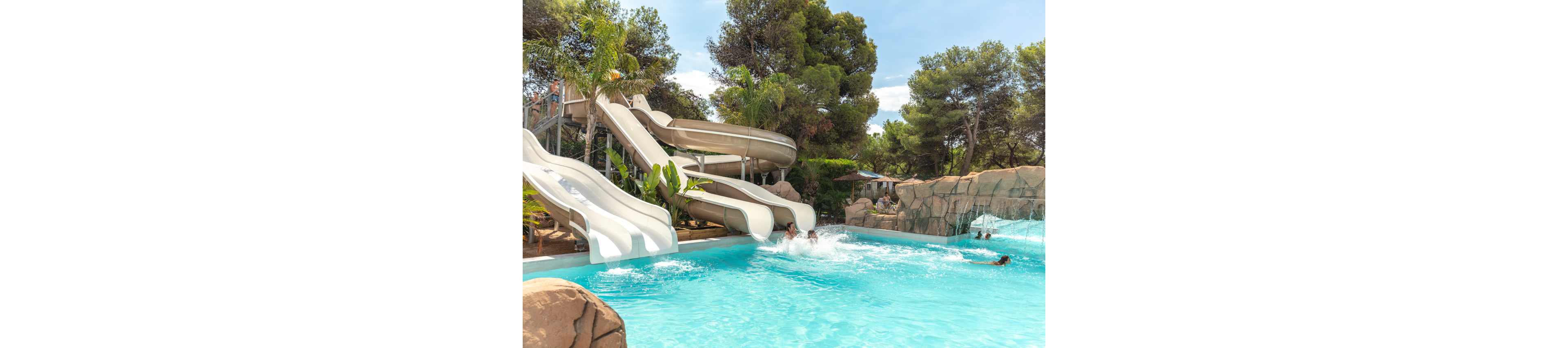 Several people slide into a bright blue swimming pool from large curving water slides. The scene is set in an outdoor water park surrounded by lush green trees and rocky landscaping. Sunlight reflects off the water, emphasizing a fun and energetic summer atmosphere. The image captures motion, splashes, and playful activity in a family-friendly setting.