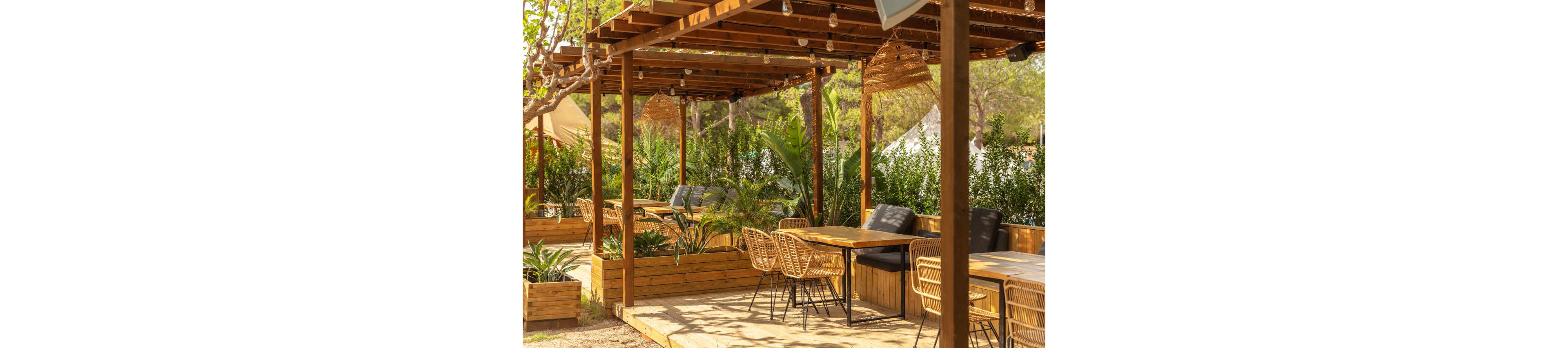 An inviting outdoor seating area features wooden pergolas and rattan chairs arranged around simple tables. Lush green plants in raised wooden planters create a natural border and add privacy to the terrace. Warm sunlight filters through the pergola beams and hanging string lights, enhancing the relaxed atmosphere. The scene suggests a comfortable café or garden lounge ideal for socializing or dining outside.