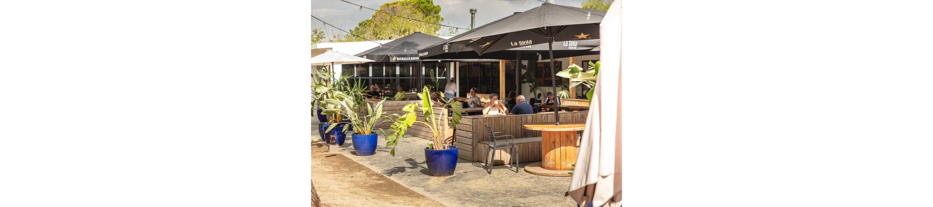 An outdoor café terrace features several groups of people seated at wooden tables under large black umbrellas. Blue planters with green tropical plants separate the seating areas and add color to the scene. The setting appears relaxed and social, with customers enjoying food and drinks in the shade. In the background, the café building and surrounding greenery are visible under a bright, sunny sky.