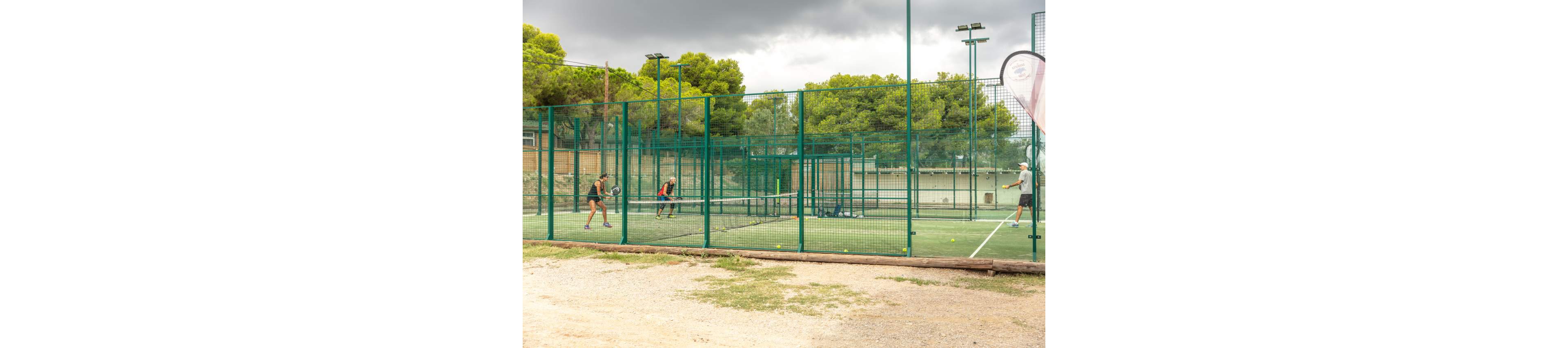 Several people play padel on an outdoor court surrounded by high green fencing. The scene is set in a sports facility with trees and buildings in the background under a cloudy sky. Players hold rackets and appear engaged in an active match. Tennis balls are scattered along the ground near the fence, emphasizing the ongoing game.