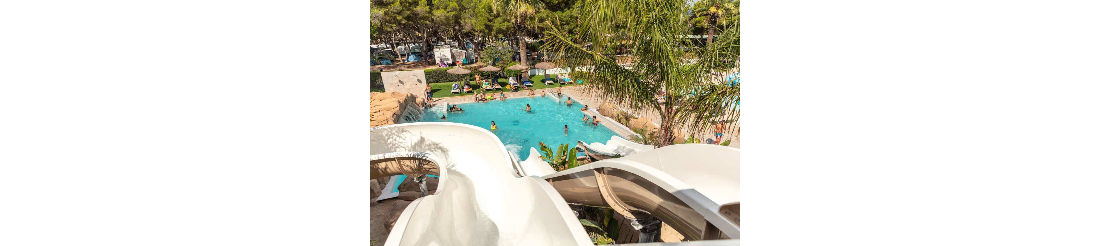 A large outdoor swimming pool is surrounded by lush palm trees and sun loungers at a resort. Several people relax in the turquoise water while others sit under straw parasols on the pool deck. Curving white waterslides dominate the foreground, leading down into the pool area. The bright daylight and greenery create a warm, summery vacation atmosphere.