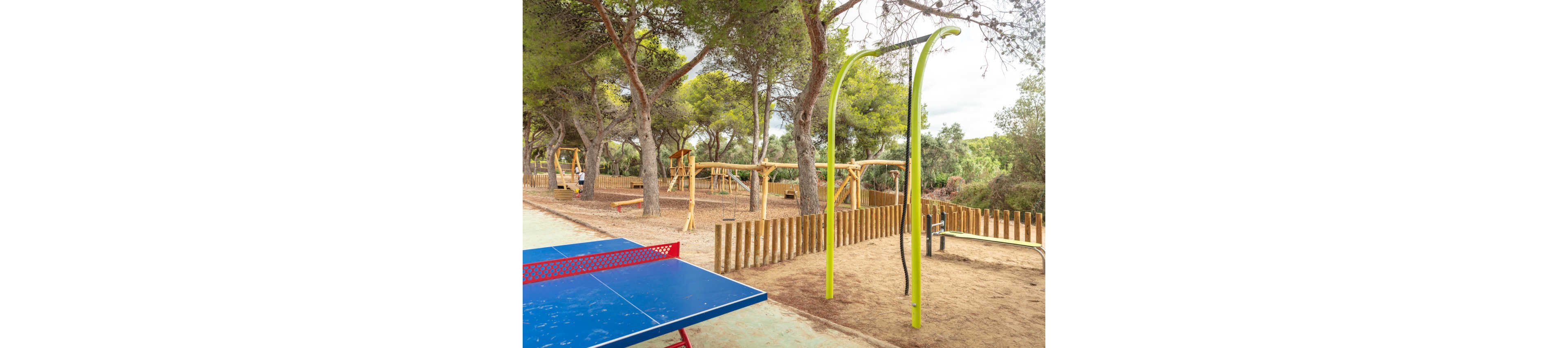 An outdoor recreation area features a blue table tennis table in the foreground. Behind it, a wooden playground with swings, climbing structures, and other equipment is set among tall pine trees. A bright green metal frame and exercise equipment add to the variety of activities available. The ground is covered with dirt and pine needles, and a wooden fence separates the play area from the path.
