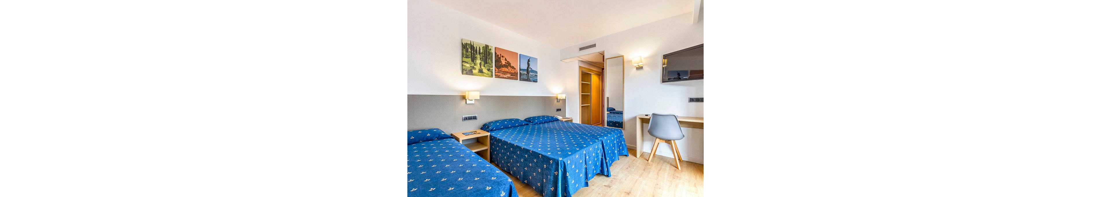 A modern hotel room features two twin beds covered with blue patterned bedspreads. The space includes light wood bedside tables, a desk with a gray chair, and a wall-mounted flat-screen television. Colorful landscape artwork decorates the wall above the beds, while wall sconces provide warm lighting. A wardrobe with mirrored door and light wood flooring complete the clean, contemporary interior.