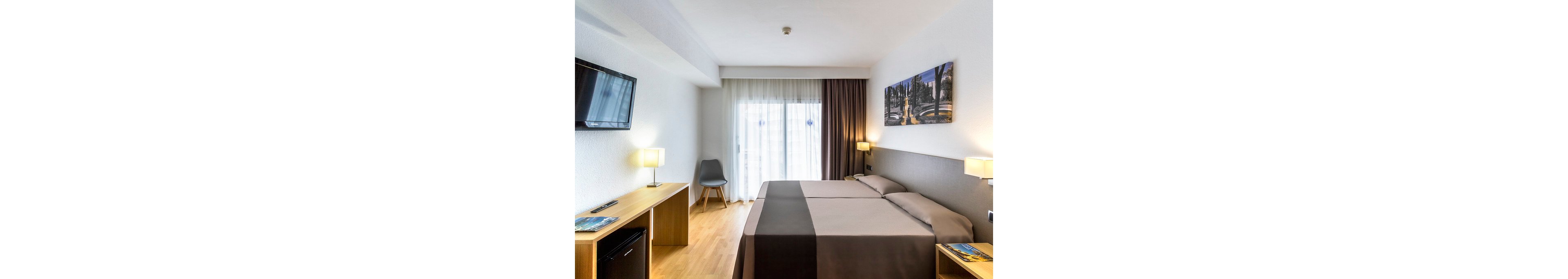 Bright contemporary hotel room featuring two twin beds pushed together with neutral bedding. The space includes a wooden desk with a chair, bedside tables, and two table lamps providing warm light. A flat-screen television is mounted on the wall opposite the beds, and a mini fridge is integrated into the desk. Large sheer curtains cover a glass door or window, allowing natural daylight to fill the room, while a framed artwork decorates the wall above the headboard.