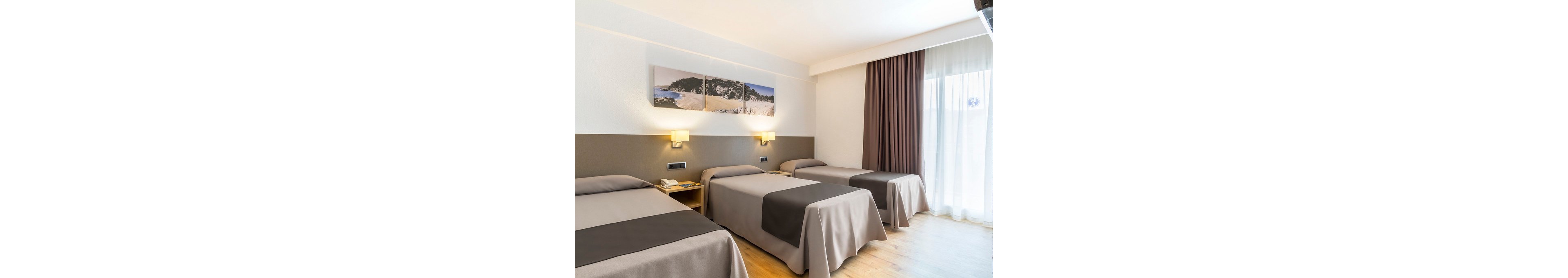 Modern hotel room featuring three neatly made single beds in neutral beige and brown tones. The space is well lit with wall-mounted lamps and natural light from a large window with sheer and blackout curtains. A small bedside table with a telephone and notepad sits between two of the beds. Above the beds, a three-panel landscape artwork adds visual interest to the clean, minimalist decor.