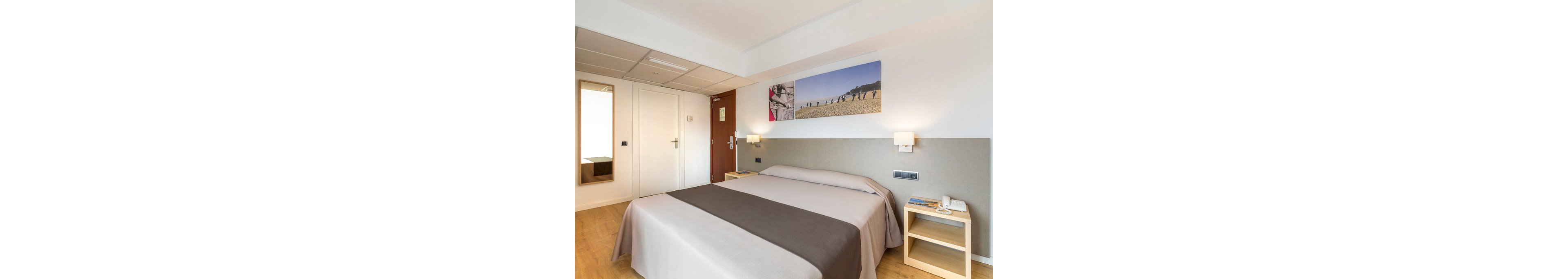 Bright contemporary hotel room featuring a neatly made double bed with neutral bedding. The space includes light wood flooring, bedside tables with reading materials and a telephone, and wall-mounted lamps. A large mirror and white closet doors enhance the sense of space, while a wooden entrance door anchors the room. A landscape photograph above the bed adds a touch of color and decor.