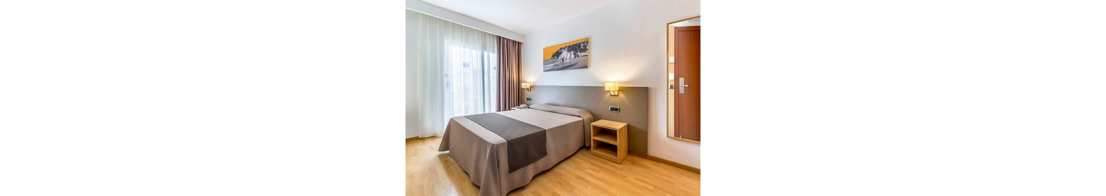 A contemporary hotel room features a neatly made double bed with neutral bedding and a contrasting dark runner. Natural light enters through large sheer curtains beside heavier drapes, illuminating the light wood floor. A small bedside table, wall-mounted lamps, and a landscape artwork above the headboard add simple decorative touches. The overall space feels clean, minimal, and inviting for guests.