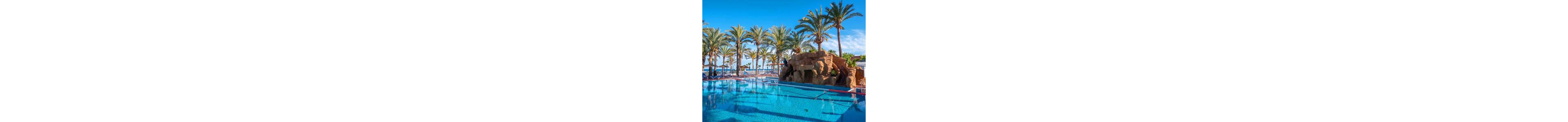 A large clear blue swimming pool reflects tall palm trees under a bright sunny sky. The pool is surrounded by a white fence and overlooks a distant ocean or sea. A rock formation with a small cave-like opening adds a playful, natural element to the scene. The overall setting suggests a tropical resort ideal for relaxation and vacation.