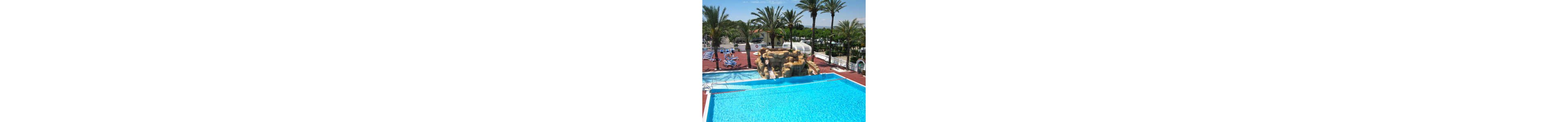A bright outdoor swimming pool glistens under clear sunlight at a resort. In the center, a rocky structure features a waterslide leading into the pool. Palm trees and sun loungers surround the pool area, creating a tropical vacation atmosphere. The scene suggests a relaxing holiday setting with recreational water activities.